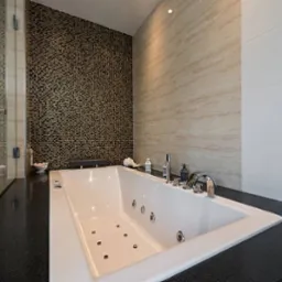 Nowoczesna łazienka z białą wanną z hydromasażem na czarnym blacie, mozaikowa ściana w odcieniach złota i brązu, oraz szklana kabina prysznicowa