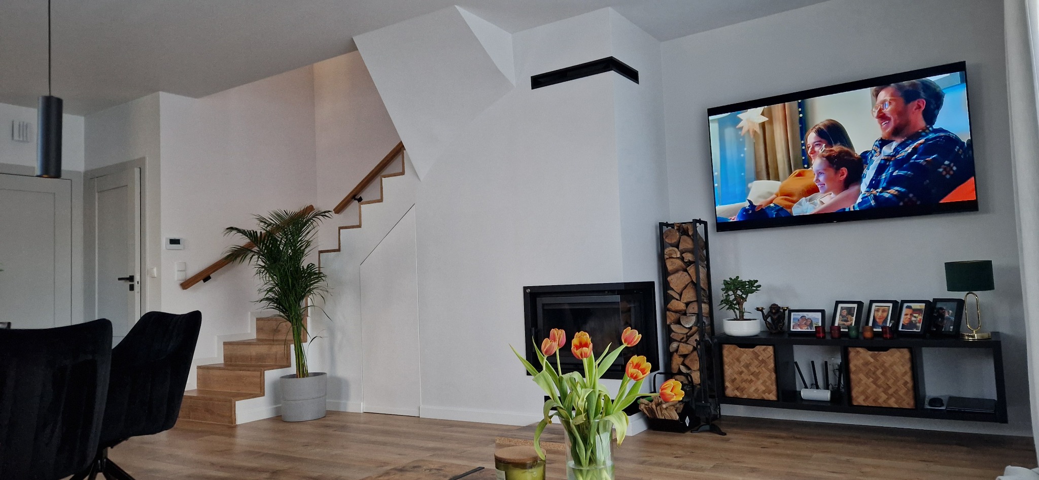 Jasne wnętrze salonu z drewnianymi schodami, kominkiem i telewizorem wyświetlającym rodzinę. Na pierwszym planie bukiet tulipanów w szklanym wazonie.