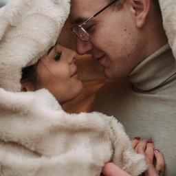 Bartosz Kwiatkowski - Zakochana para przytulona pod miękkim, futrzanym okryciem, z bliska widoczne profile twarzy, ona z czerwonym manicure i tatuażem krzyżyka na dłoni, on w okularach i golfie.