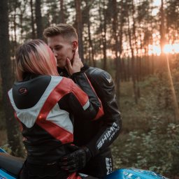 Bartosz Kwiatkowski - Para w skórzanych strojach motocyklowych, obejmująca się na turkusowym motocyklu w lesie, z promieniami zachodzącego słońca w tle.