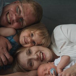 Bartosz Kwiatkowski - Szczęśliwa rodzina leżąca na szarym tle: uśmiechnięty ojciec w okularach, córka w niebieskiej sukience, mama i śpiący noworodek z niebieskim smoczkiem.