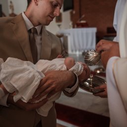 Bartosz Kwiatkowski - Ojciec w beżowym garniturze trzyma dziecko w białej sukience podczas chrztu, w tle fragment wnętrza kościoła i ksiądz polewający wodą.