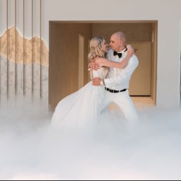 Bartosz Kwiatkowski - Uśmiechnięta para w białych, eleganckich strojach, obejmująca się w kłębach niskiego dymu. Kobieta w sukni, mężczyzna w koszuli i muszce, na tle nowoczesnej ściany z podświetlanym wzorem gór...