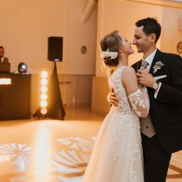 Bartosz Kwiatkowski - Młoda para w sukni ślubnej z koronkowymi rękawami i garniturze tańczy, patrząc sobie czule w oczy. W tle DJ z oświetleniem i wzory światła na parkiecie.