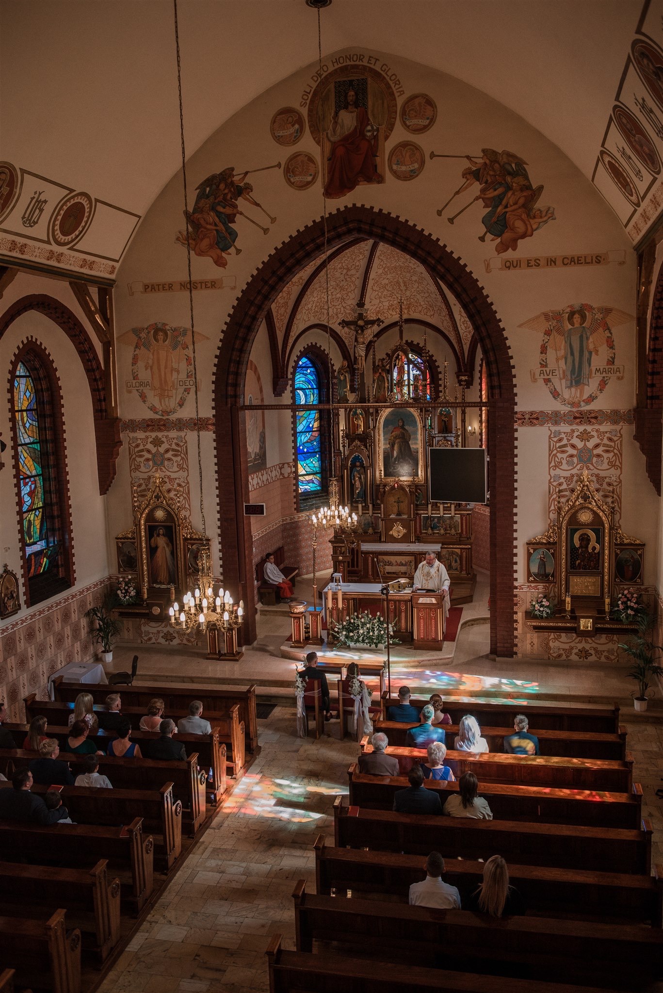 Wnętrze kościoła podczas ceremonii, widoczne ławki z gośćmi, ołtarz z duchownym, witraże i dekoracje malarskie na ścianach i suficie.