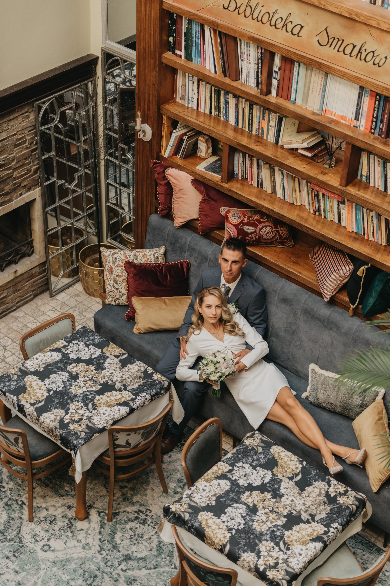 Para młoda siedzi na szarej sofie w restauracji z biblioteczką w tle. Widok z góry na stoliki z kwiecistymi obrusami.