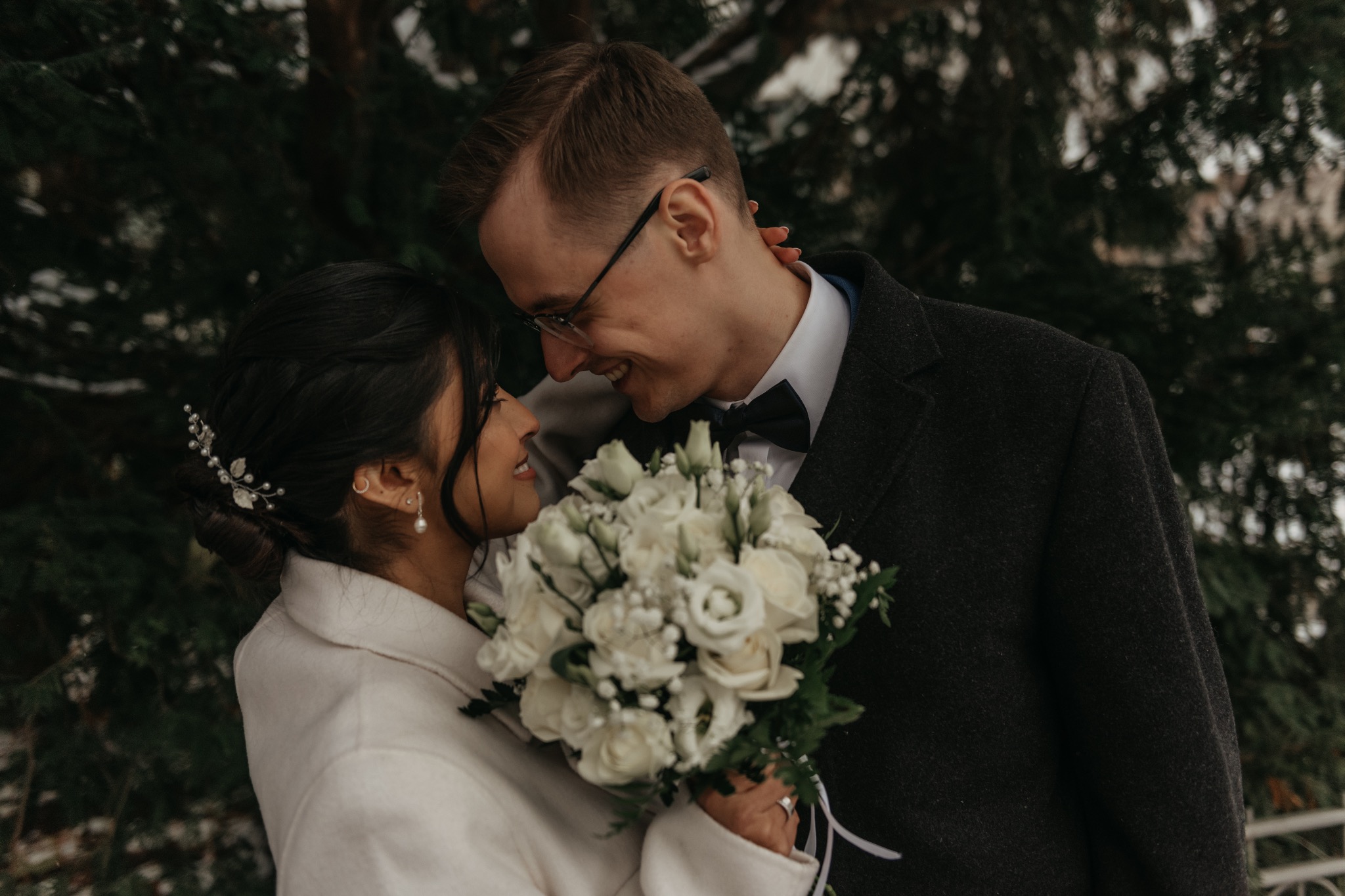 Uśmiechnięta para młoda w zimowej scenerii, panna młoda z bukietem białych róż, pan młody w muszce i okularach, delikatne oświetlenie.