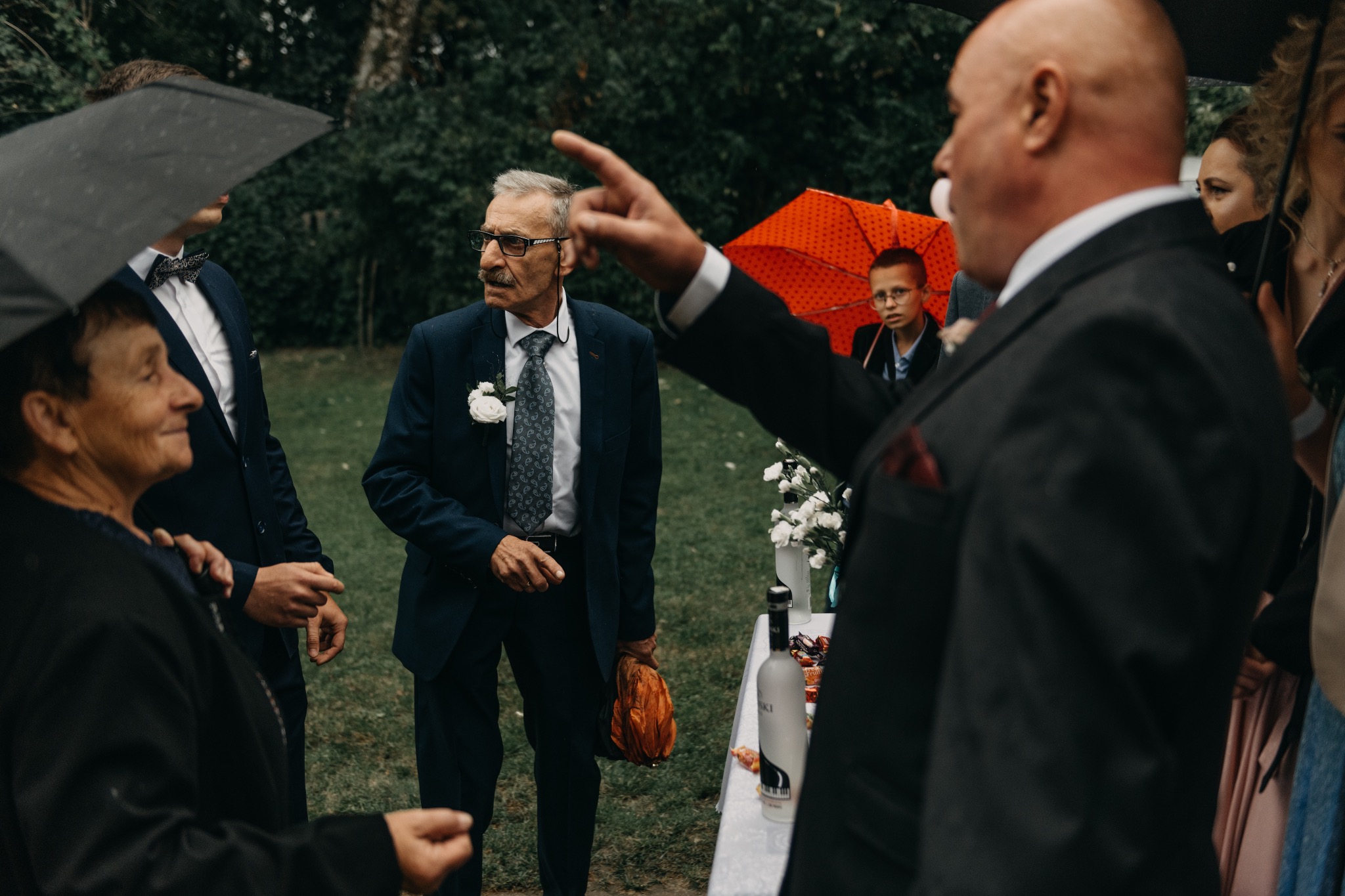 Goście weselni w eleganckich strojach pod parasolami podczas przyjęcia na świeżym powietrzu, w tle stół z przekąskami i napojami.