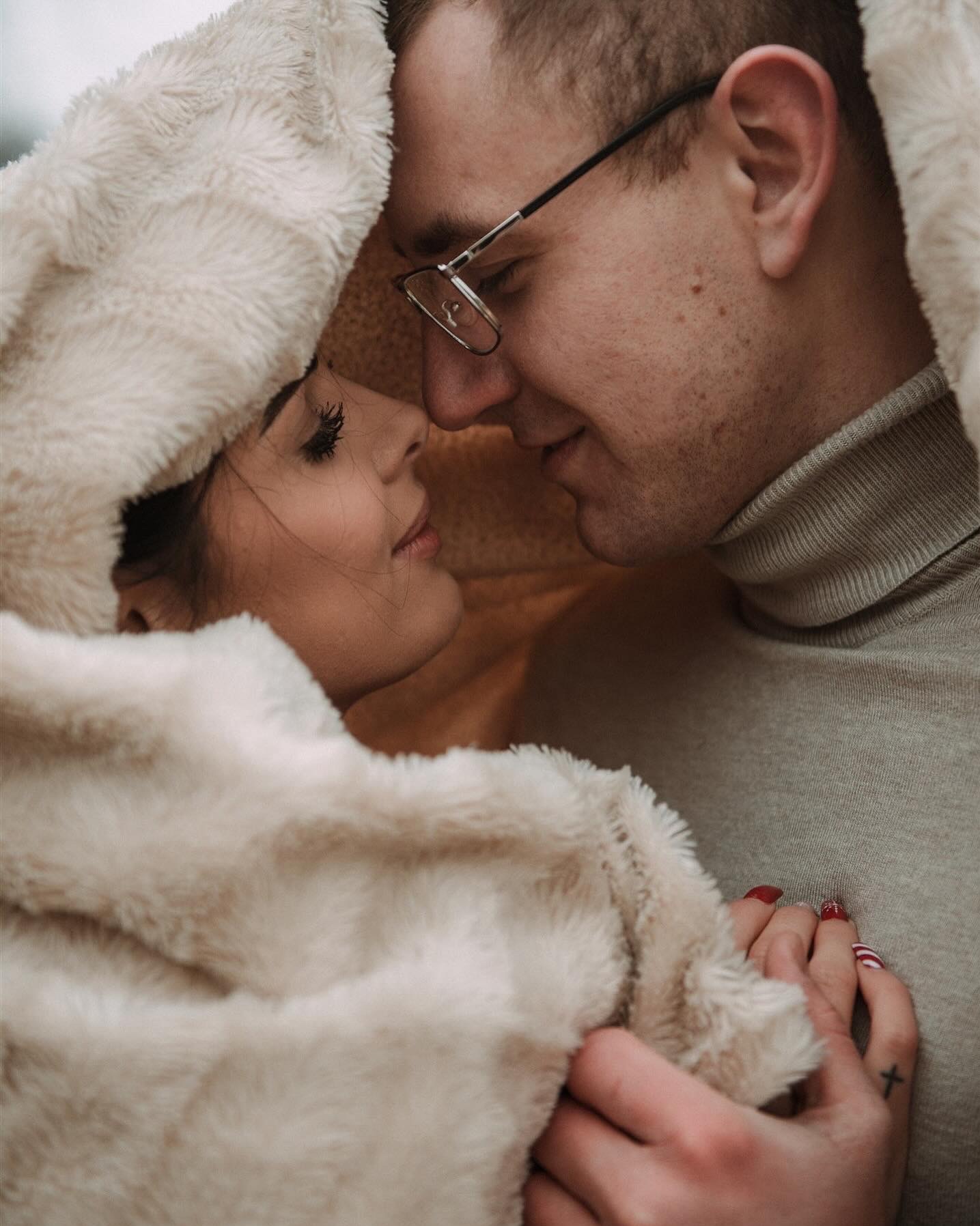 Zakochana para przytulona pod miękkim, futrzanym okryciem, z bliska widoczne profile twarzy, ona z czerwonym manicure i tatuażem krzyżyka na dłoni, on w okularach i golfie.