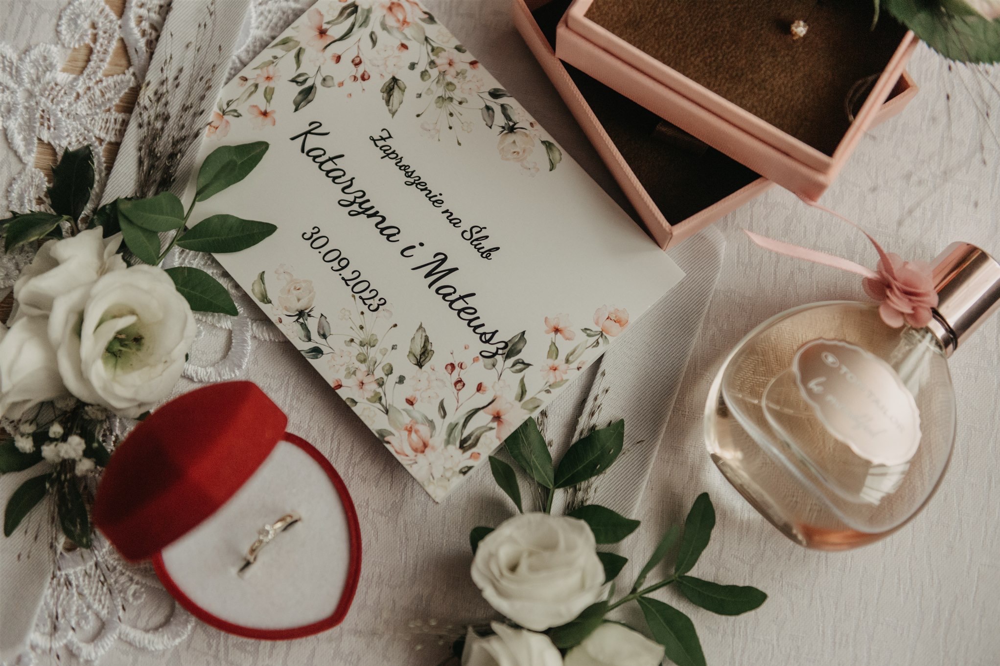 Zaproszenie ślubne z datą 30.09.2023, pierścionek zaręczynowy w czerwonym pudełku w kształcie serca, perfumy i pudełko na obrączki na białym obrusie.