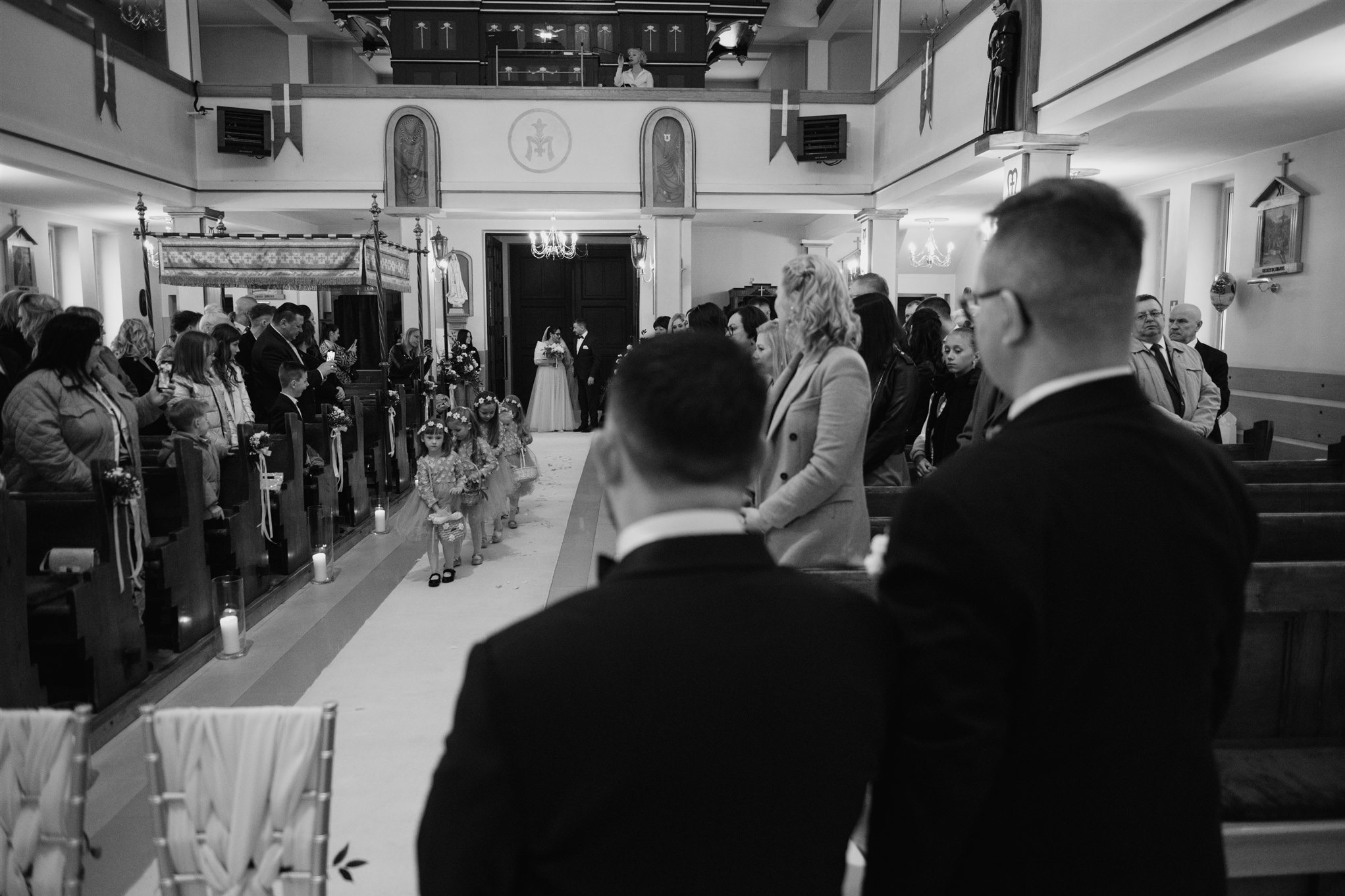 Czarno-białe zdjęcie z wnętrza kościoła podczas ceremonii ślubnej, z perspektywy gościa za parą młodą i dziewczynkami sypiącymi kwiaty, z widocznym balkonem dla orkiestry.
