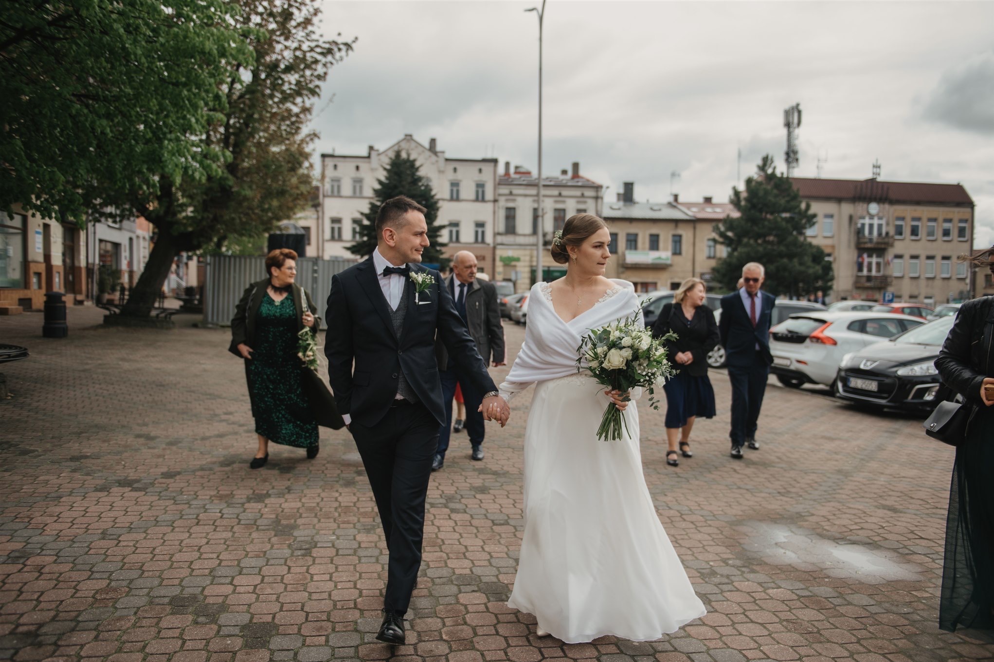 Para młoda, trzymając się za ręce, idzie po brukowanym placu w otoczeniu gości weselnych, w tle budynki miejskie i zaparkowane samochody.