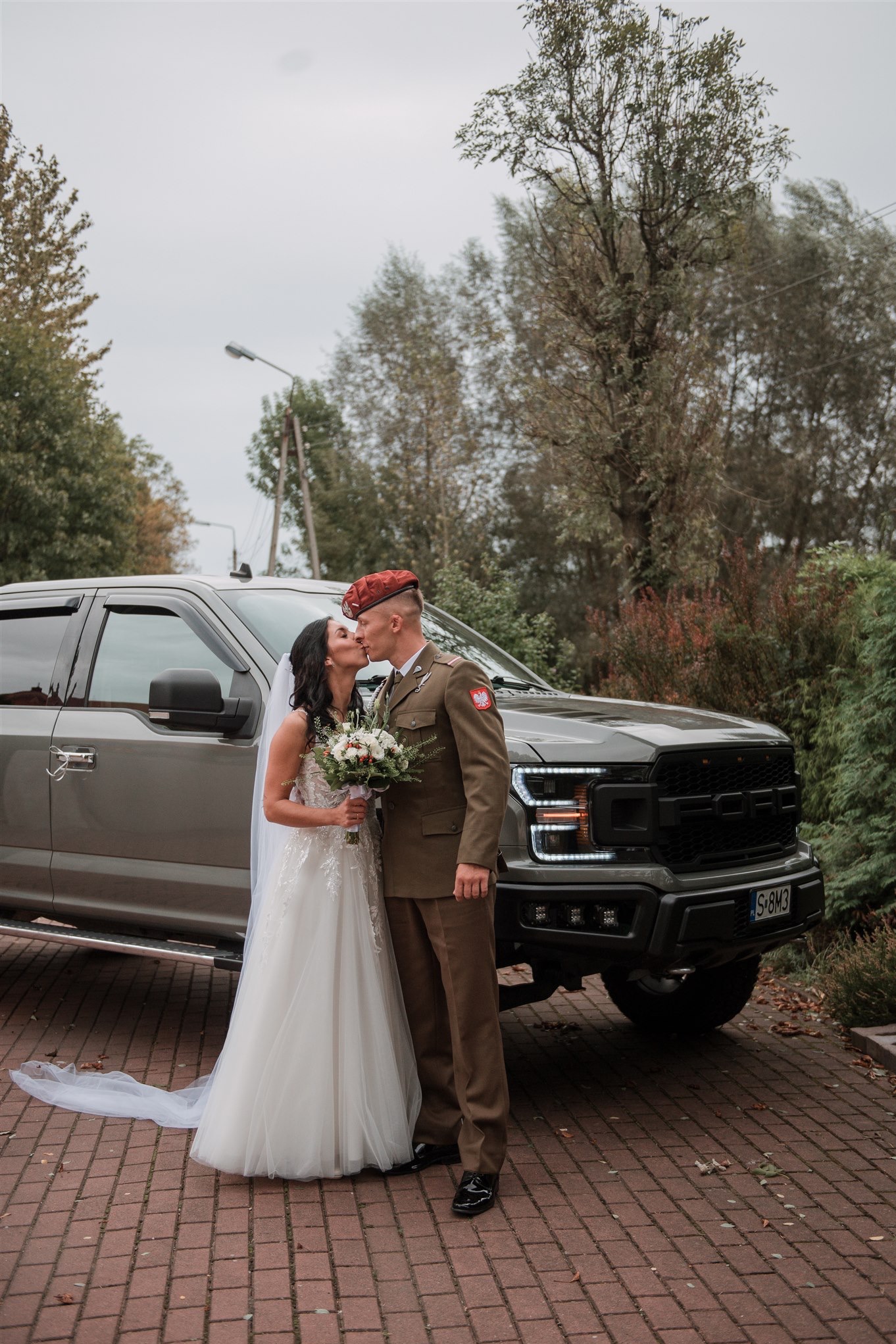 Pocałunek nowożeńców na tle szarego pickupa Ford Raptor; panna młoda w białej sukni z bukietem, pan młody w mundurze Wojska Polskiego i czerwonym berecie.