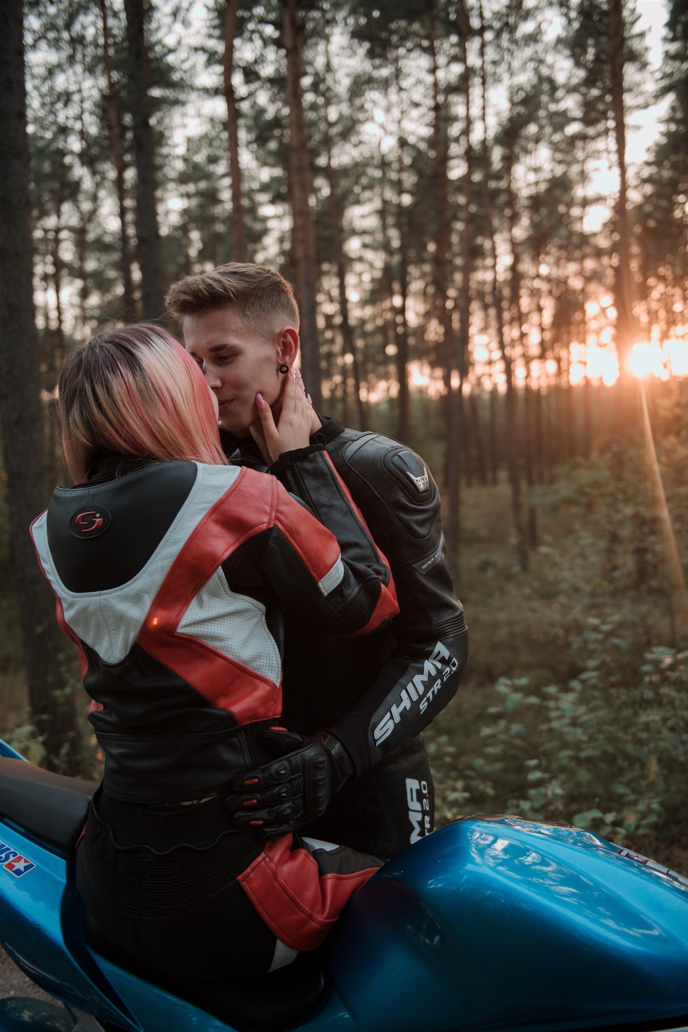 Para w skórzanych strojach motocyklowych, obejmująca się na turkusowym motocyklu w lesie, z promieniami zachodzącego słońca w tle.