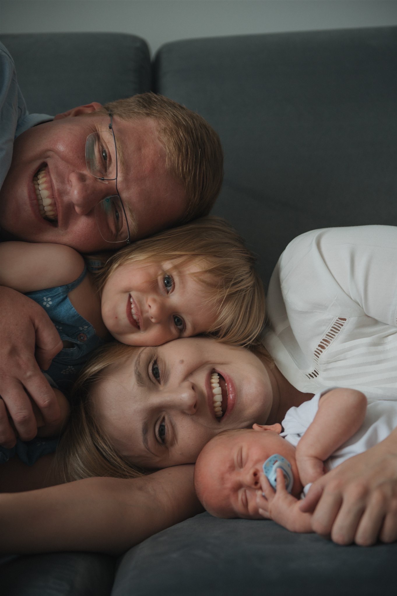 Szczęśliwa rodzina leżąca na szarym tle: uśmiechnięty ojciec w okularach, córka w niebieskiej sukience, mama i śpiący noworodek z niebieskim smoczkiem.