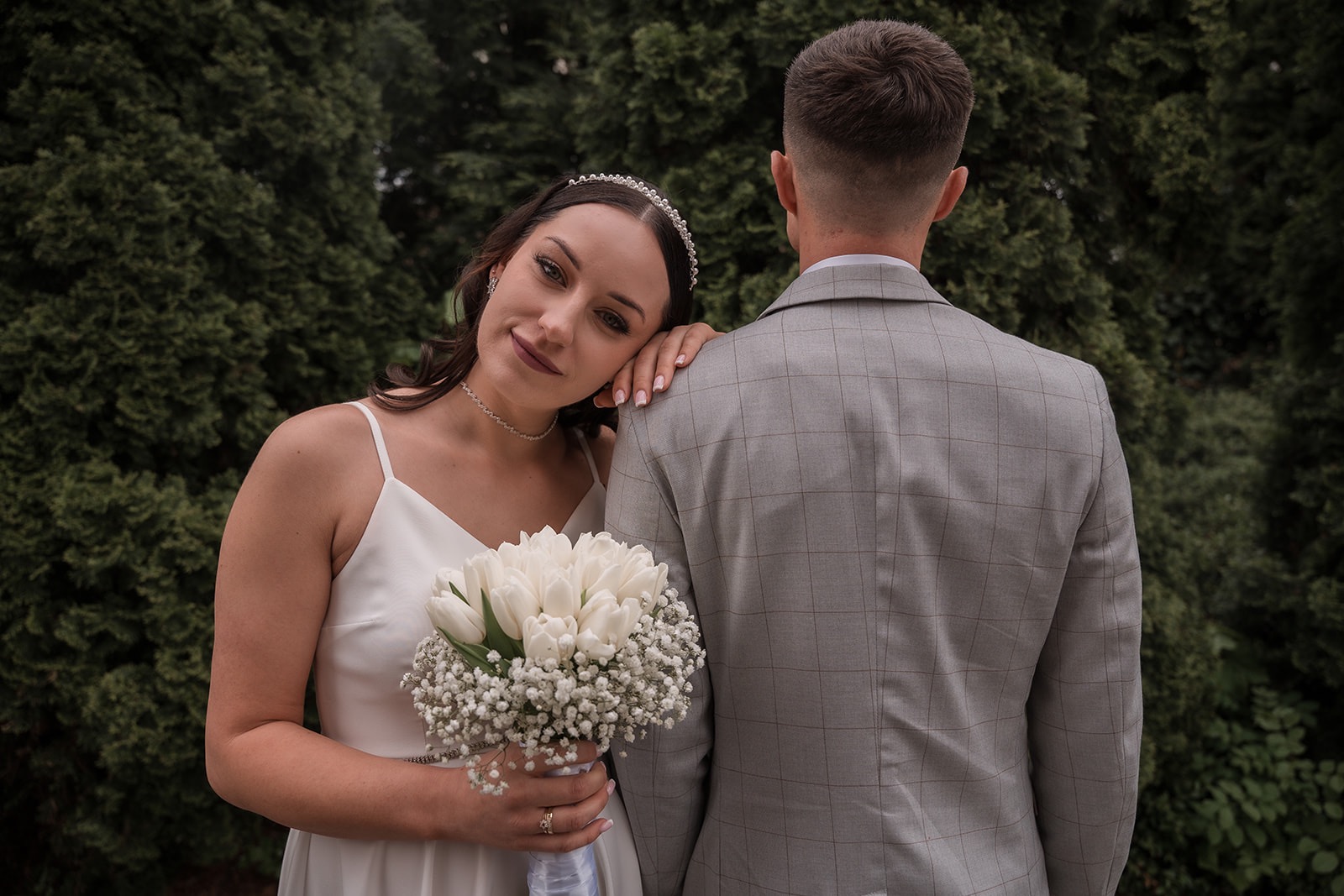 Panna młoda z bukietem białych tulipanów opiera się o ramię pana młodego w szarej marynarce. Tło z ciemnozielonych drzew. Delikatny uśmiech, subtelna biżuteria.