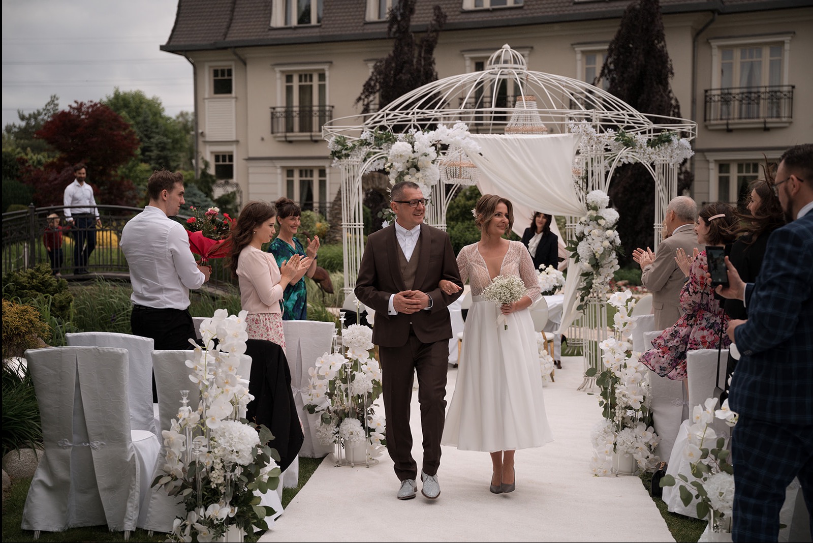 Para młoda idzie udekorowaną ścieżką, otoczona gośćmi i kwiatami, w tle biała altana i budynek. Pan młody w brązowym garniturze, panna młoda w białej sukni.