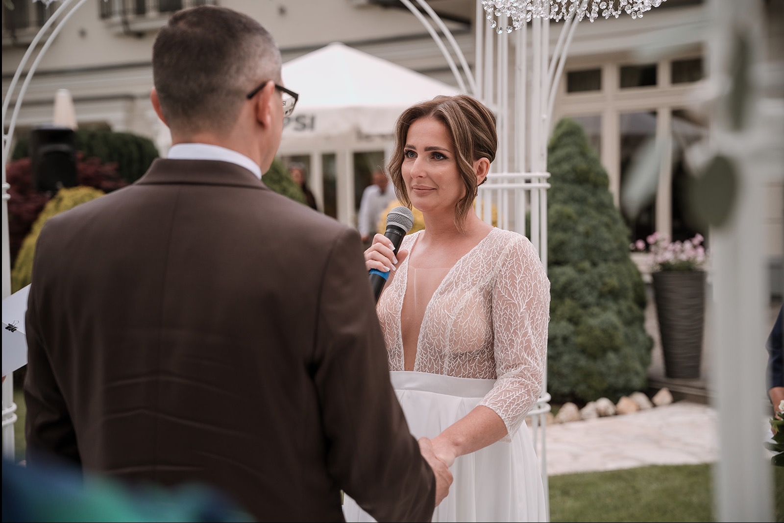 Panna młoda z mikrofonem podczas ceremonii ślubnej, w tle biała altana i mężczyzna w brązowym garniturze. Ujęcie z bliska, skupione na wyrazie twarzy kobiety.