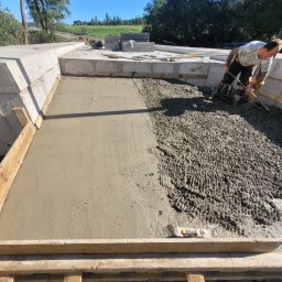 KIRBUD Krzysztof Kirkiewicz - Pracownik w gumowych butach rozprowadza świeży beton w szalunku fundamentowym, część powierzchni już wyrównana, widoczne zbrojenie i narzędzia.