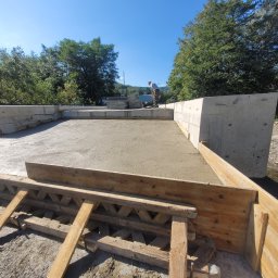 KIRBUD Krzysztof Kirkiewicz - Świeżo wylana betonowa płyta z drewnianym szalunkiem, widoczny pracownik wygładzający powierzchnię, w tle drzewa i błękitne niebo.