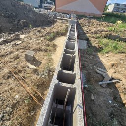 KIRBUD Krzysztof Kirkiewicz - Betonowe bloczki fundamentowe zbrojone stalą, ułożone w wykopie, z widoczną czerwoną linią poziomowania, na tle placu budowy z hałdą ziemi i budynkiem mieszkalnym.