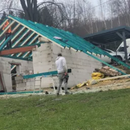 Wznoszenie ścian i konstrukcji dachu domu murowanego z betonu komórkowego, widoczni dwaj pracownicy na dachu, jeden macha ręką, w tle las i częściowo widoczny samochód dostawczy.