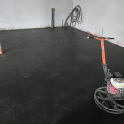 Świeżo wylana, mokra posadzka betonowa wewnątrz pomieszczenia, z pomarańczowymi znacznikami poziomów przy ścianach i maszyną do zacierania betonu z pomarańczową rączką. W tle widoczne wystające...