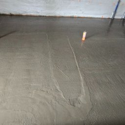 KIRBUD Krzysztof Kirkiewicz - Świeżo wylana, jeszcze mokra, szara wylewka podłogowa z wystającymi rurkami instalacyjnymi, zabezpieczona folią przy ścianach.