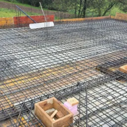 Zbrojenie płyty fundamentowej z siatki stalowej, drewniane szalunki obwodowe i skrzynki na tle zieleni.