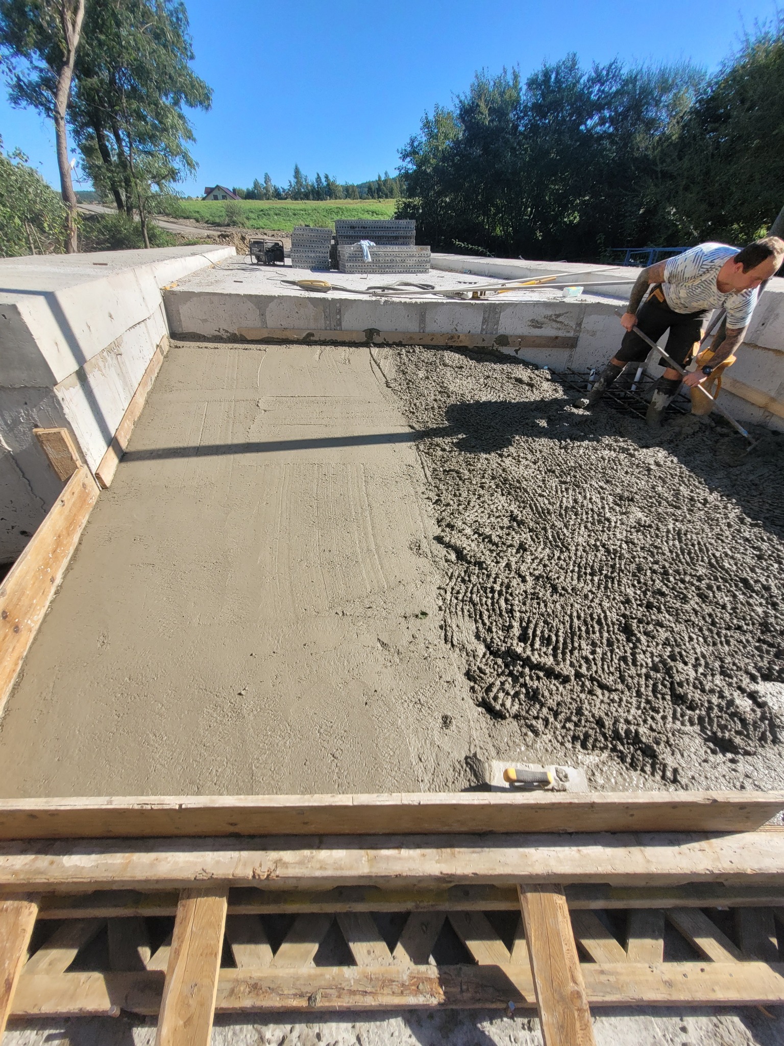 Pracownik w gumowych butach rozprowadza świeży beton w szalunku fundamentowym, część powierzchni już wyrównana, widoczne zbrojenie i narzędzia.