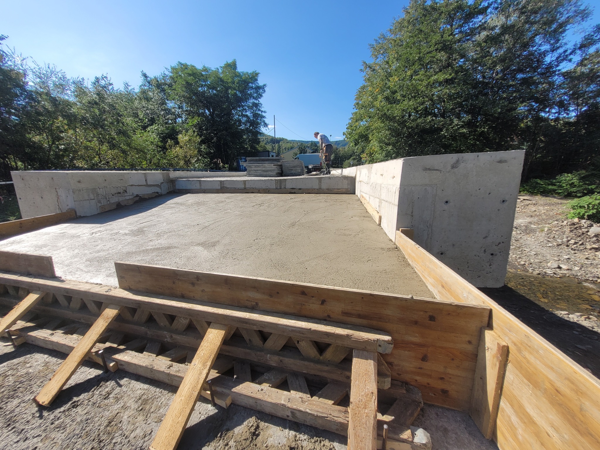 Świeżo wylana betonowa płyta z drewnianym szalunkiem, widoczny pracownik wygładzający powierzchnię, w tle drzewa i błękitne niebo.