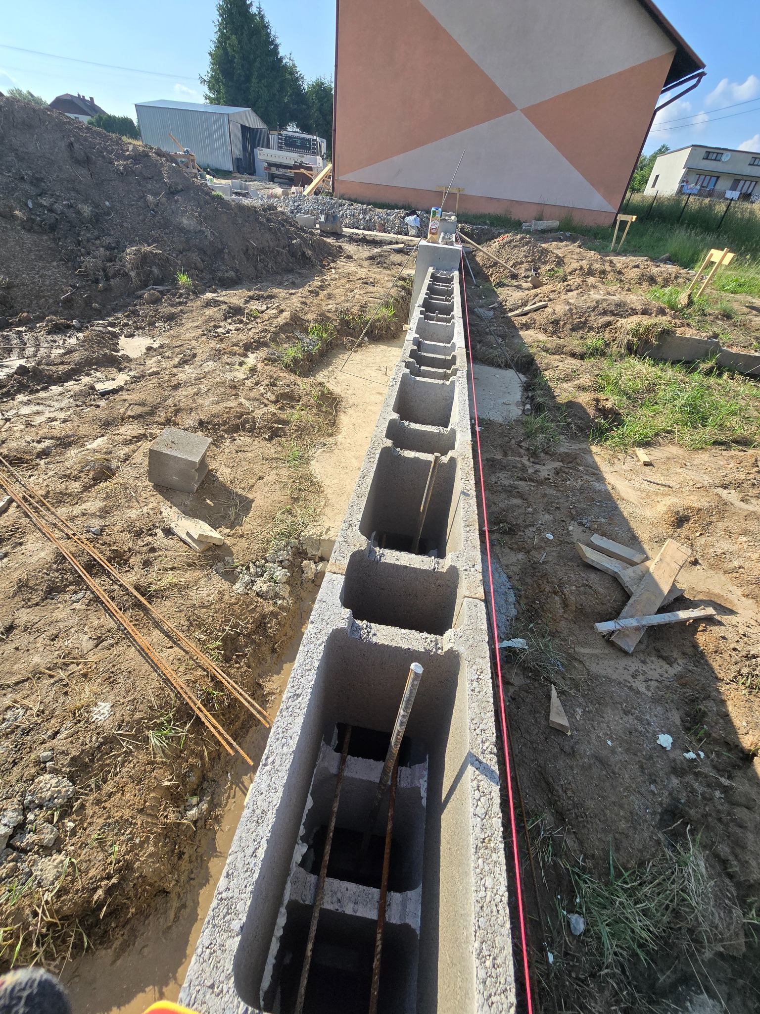 Betonowe bloczki fundamentowe zbrojone stalą, ułożone w wykopie, z widoczną czerwoną linią poziomowania, na tle placu budowy z hałdą ziemi i budynkiem mieszkalnym.