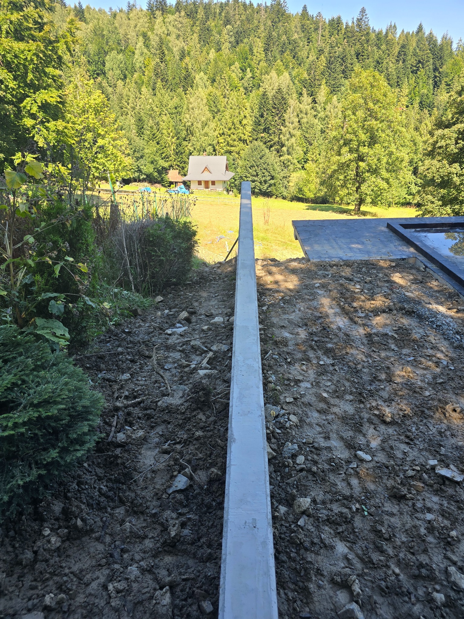 Szczegółowy widok szalunku fundamentowego z betonu, przygotowanego pod wylewkę, na tle zieleni i krajobrazu z domem w oddali.