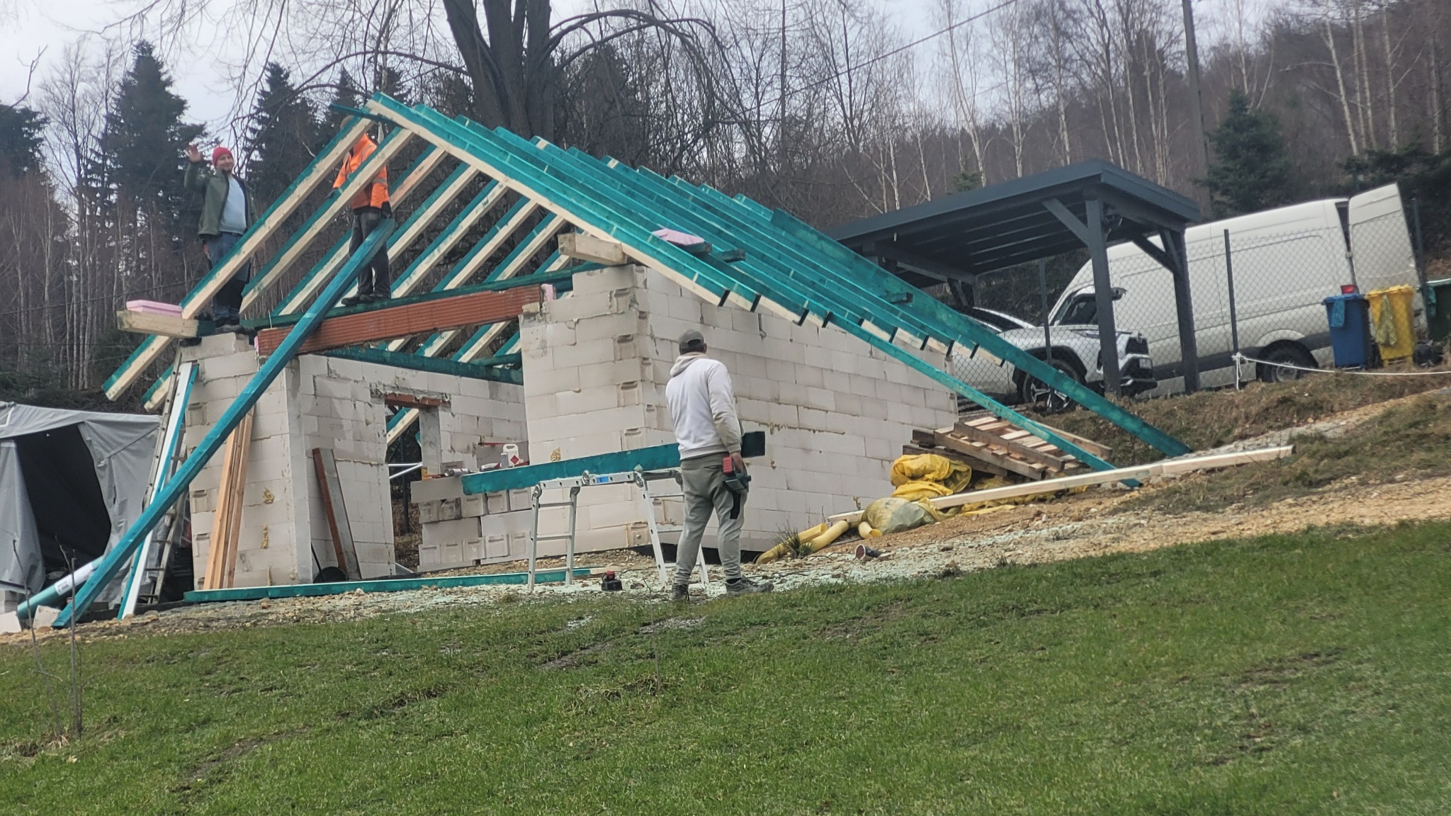 Wznoszenie ścian i konstrukcji dachu domu murowanego z betonu komórkowego, widoczni dwaj pracownicy na dachu, jeden macha ręką, w tle las i częściowo widoczny samochód dostawczy.