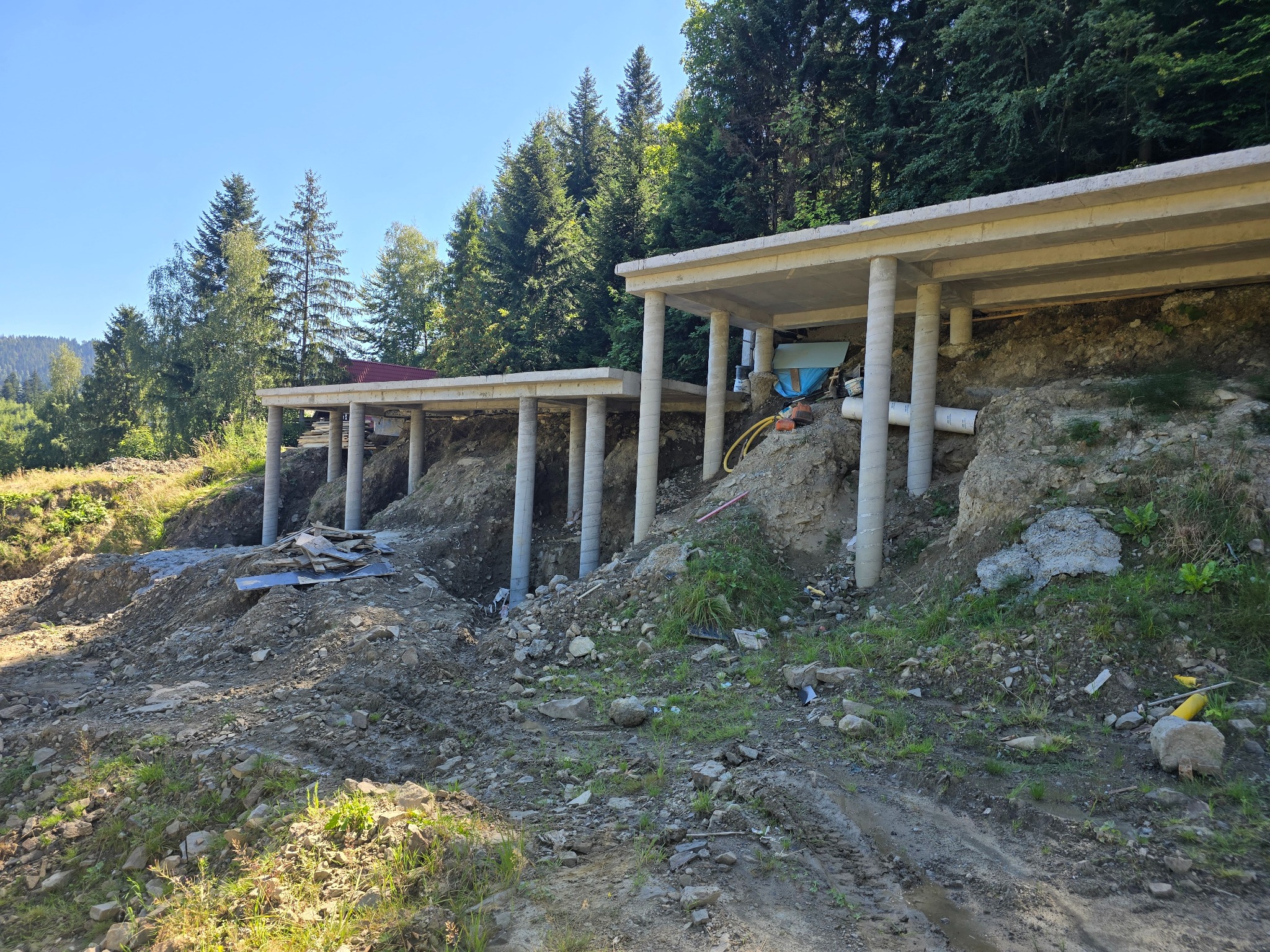 Surowe betonowe fundamenty pod budynek na nierównym terenie z widokiem na las w tle, widoczne szalunki i podpory.