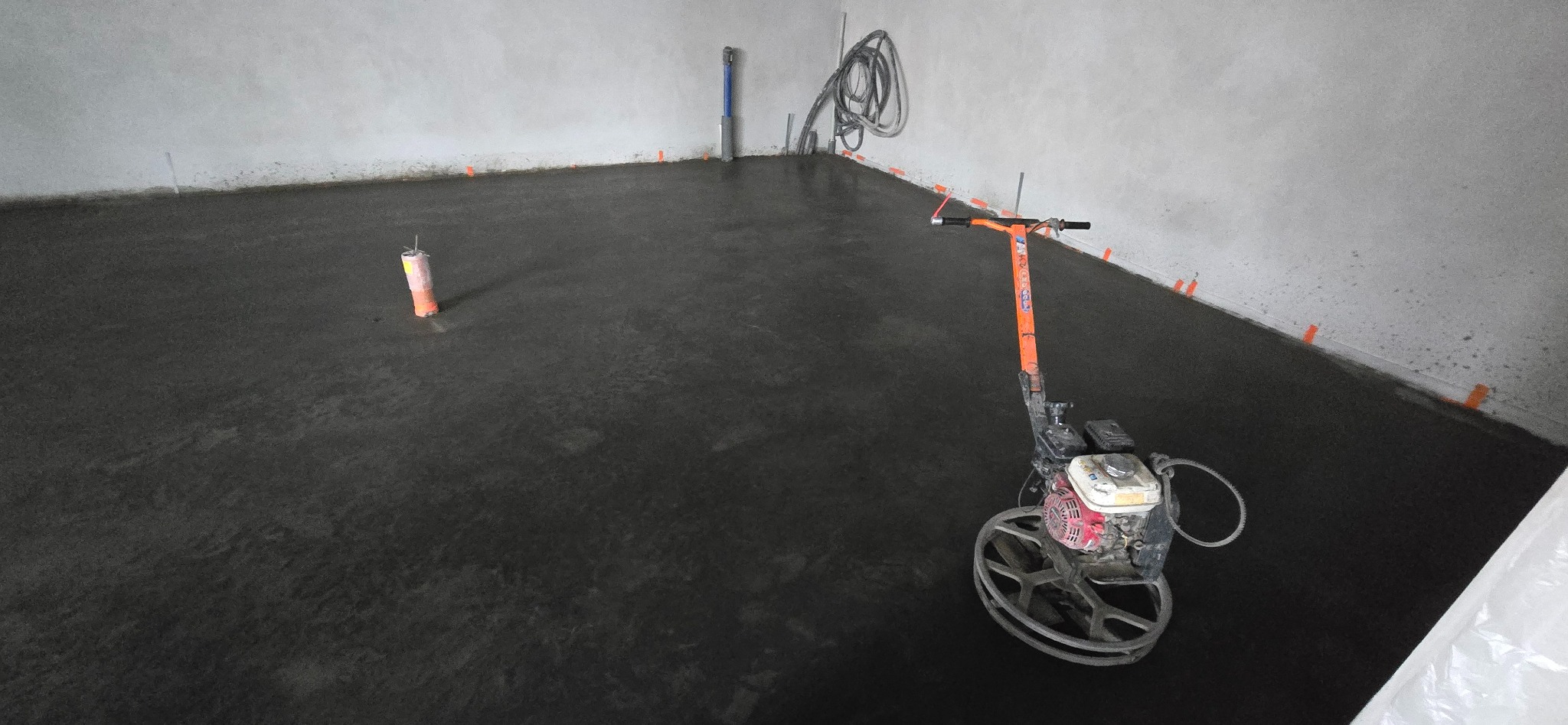 Świeżo wylana, mokra posadzka betonowa wewnątrz pomieszczenia, z pomarańczowymi znacznikami poziomów przy ścianach i maszyną do zacierania betonu z pomarańczową rączką. W tle widoczne wystające...