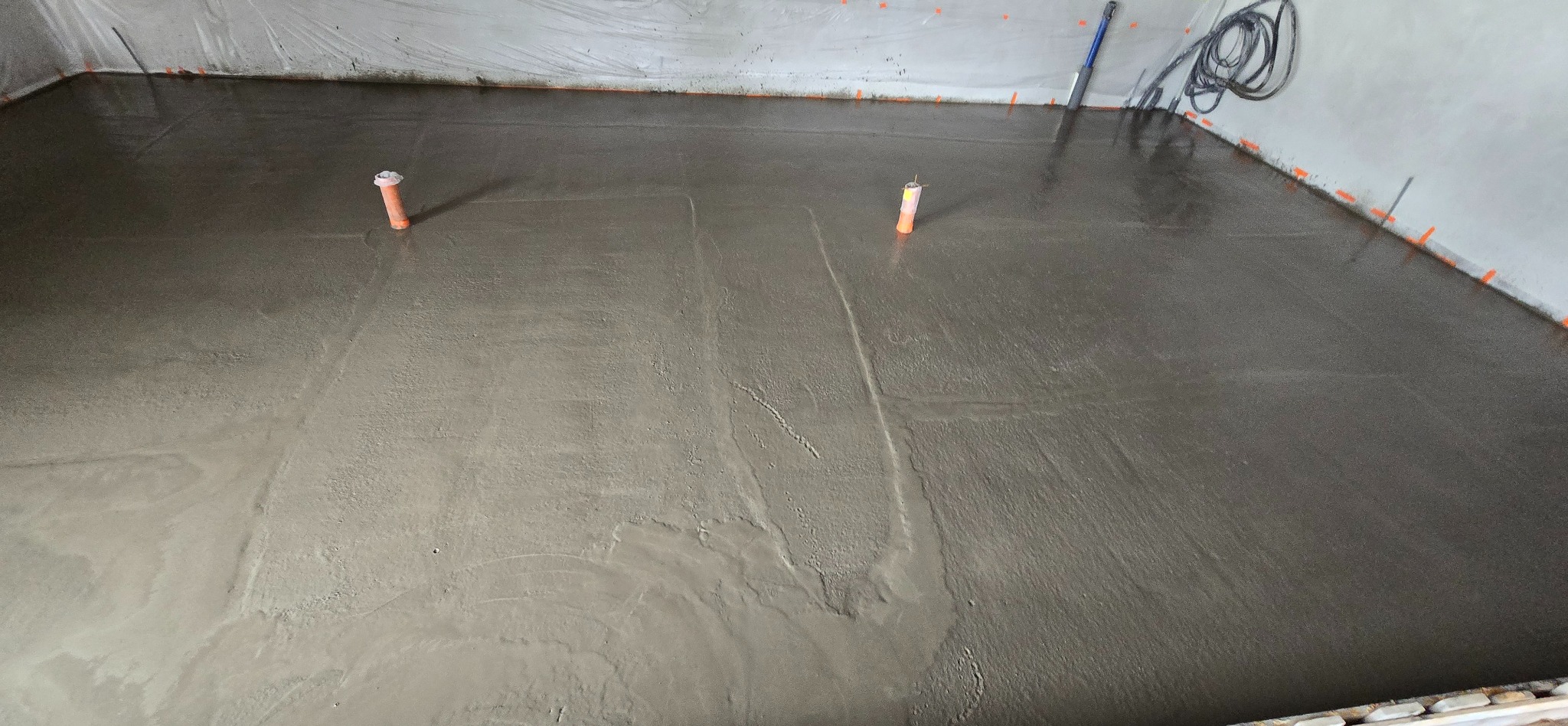 Świeżo wylana, jeszcze mokra, szara wylewka podłogowa z wystającymi rurkami instalacyjnymi, zabezpieczona folią przy ścianach.