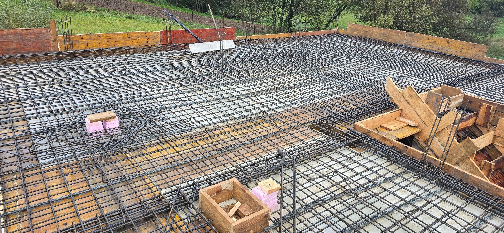 Zbrojenie płyty fundamentowej z siatki stalowej, drewniane szalunki obwodowe i skrzynki na tle zieleni.