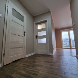 D&D USŁUGI REMONTOWO-BUDOWLANE PAWEŁ DUBIEL - Korytarz po remoncie z dwoma parami drzwi wewnętrznych z przeszkleniami, panelami podłogowymi imitującymi drewno, szarą kolorystyką ścian i widokiem na zewnątrz przez okno balkonowe.