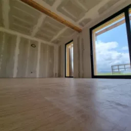 Wnętrze w trakcie remontu: ściany pokryte płytami kartonowo-gipsowymi z widocznymi łączeniami, drewniana belka stropowa, panele podłogowe, duże okno z widokiem na zewnątrz.