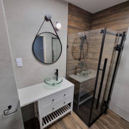 D&D USŁUGI REMONTOWO-BUDOWLANE PAWEŁ DUBIEL - Nowoczesna łazienka z kabiną prysznicową w czarnej ramie, umywalką nablatową ze szkła na białej szafce z szufladami i okrągłym lustrem zawieszonym na skórzanym pasku.