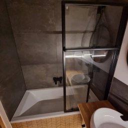 D&D USŁUGI REMONTOWO-BUDOWLANE PAWEŁ DUBIEL - Nowoczesna łazienka z wanną obudowaną drewnopodobnymi płytkami ułożonymi w jodełkę, czarna armatura i szklany parawan nawannowy z czarną ramą, umywalka nablatowa na drewnianej szafce.