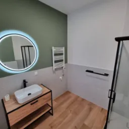 Nowoczesna łazienka z umywalką nablatową na drewnianej szafce z czarnymi elementami, okrągłe lustro z podświetleniem LED, grzejnik drabinkowy i kabina prysznicowa z czarnymi profilami.