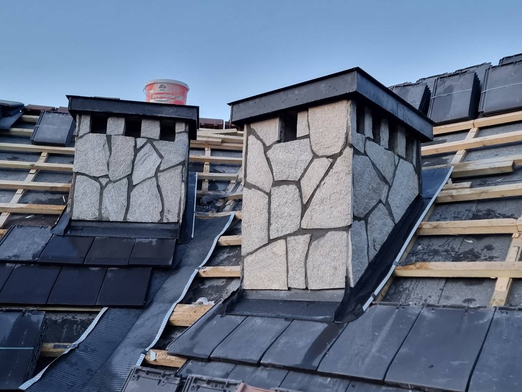 Dwa kominy wykończone nieregularnym kamieniem elewacyjnym na dachu z czarnej dachówki, widoczna konstrukcja drewniana i wiadro farby na jednym z kominów.