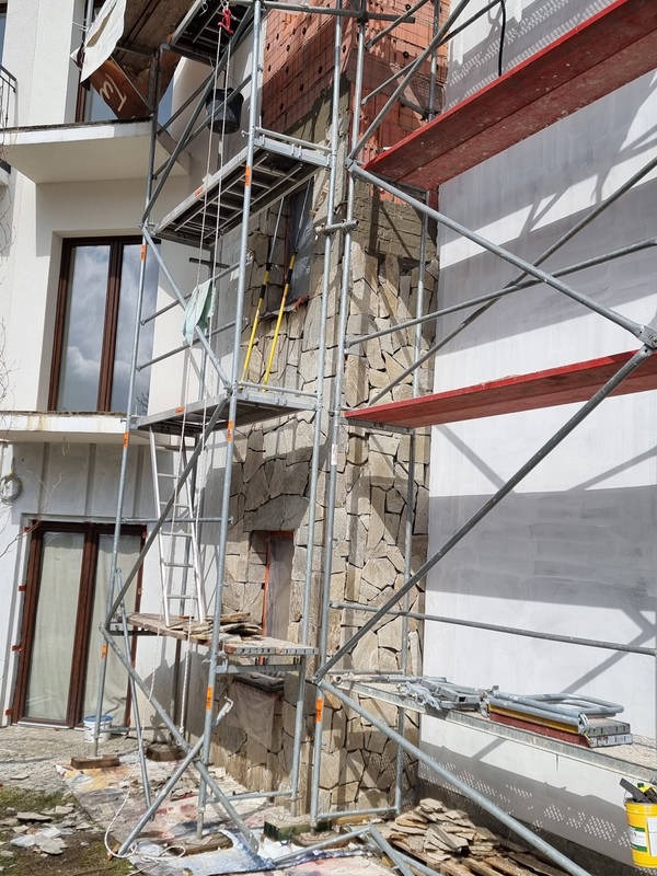 Elewacja budynku w trakcie prac wykończeniowych z użyciem kamienia naturalnego, widoczne rusztowania i materiały budowlane na ziemi.