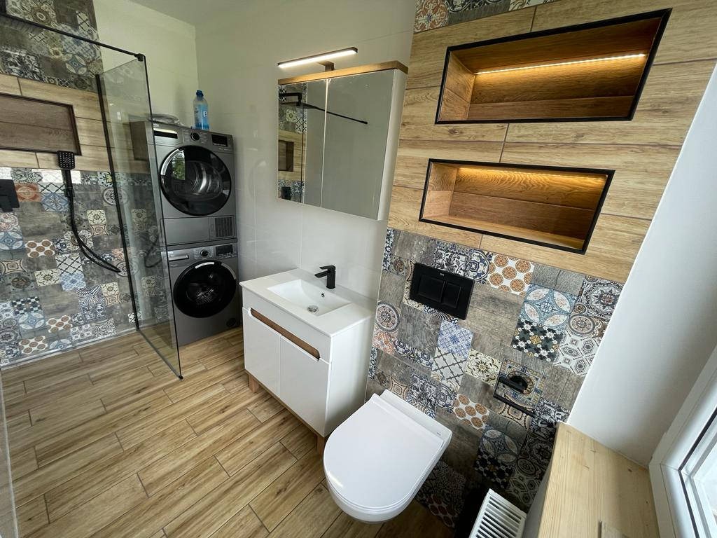 Nowoczesna łazienka z prysznicem typu walk-in, pralko-suszarką w zabudowie, umywalką nablatową na szafce i podwieszaną miską WC, wykończona płytkami patchworkowymi i drewnopodobnymi,...