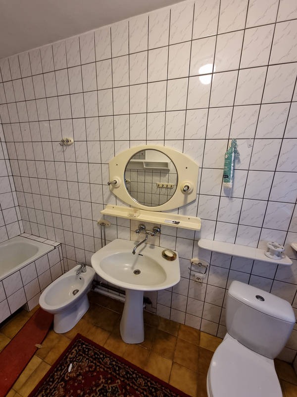 Umeblowana łazienka z białą armaturą: umywalka na postumencie, bidet, toaleta, wanna. Ściany wyłożone kwadratowymi, białymi płytkami z szarymi fugami. Nad umywalką owalne lustro z półką.