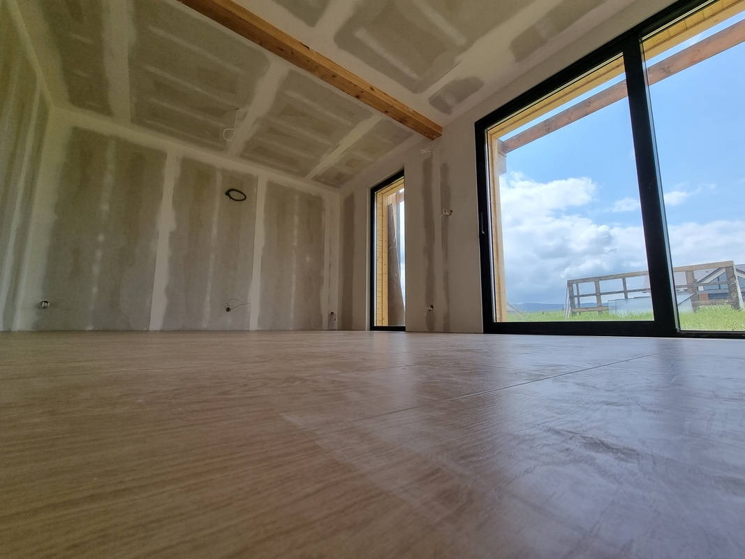 Wnętrze w trakcie remontu: ściany pokryte płytami kartonowo-gipsowymi z widocznymi łączeniami, drewniana belka stropowa, panele podłogowe, duże okno z widokiem na zewnątrz.