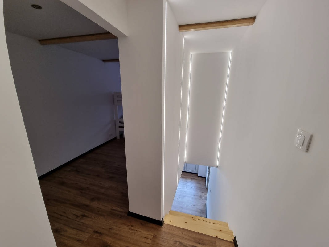 Białe wnętrze domu z drewnianymi akcentami: widok na schody w dół i pokój z łóżkiem piętrowym w tle, oświetlenie LED wzdłuż krawędzi ścian.