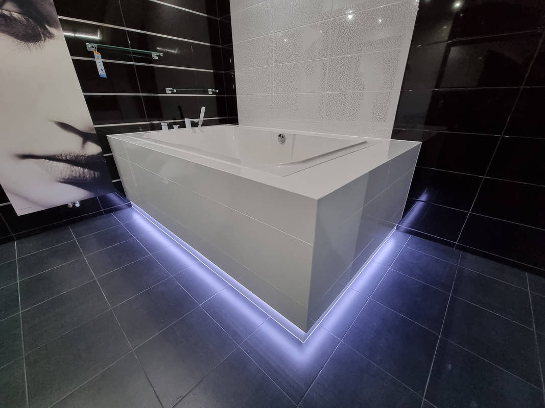 Nowoczesna, biała wanna obudowana płytkami z podświetleniem LED w dolnej części, czarne i białe płytki na ścianach, minimalistyczny design łazienki z akcentem na oświetlenie i duży plakat z twarzą...